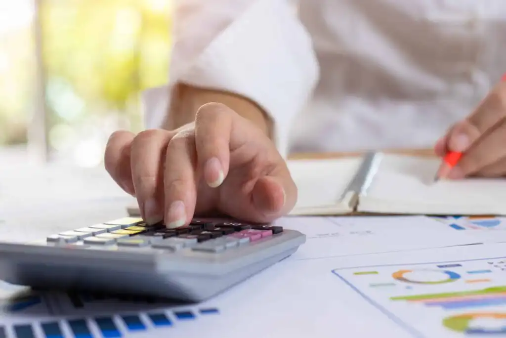 A person uses a calculator with one hand while writing in a notebook with the other. Financial charts and documents are spread out on the desk.